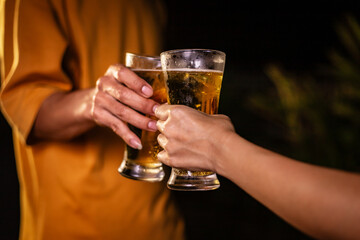 A man's hand and a woman's hand hold beer glasses in a cozy, lively evening party setting. Family and friends gather under the night sky, sharing joyful moments and laughter.