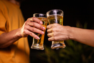 A man's hand and a woman's hand hold beer glasses in a cozy, lively evening party setting. Family and friends gather under the night sky, sharing joyful moments and laughter.