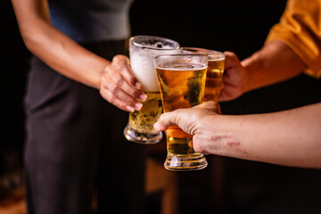 A man's hand and a woman's hand hold beer glasses in a cozy, lively evening party setting. Family and friends gather under the night sky, sharing joyful moments and laughter.