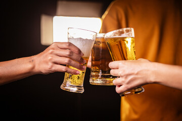 A man's hand and a woman's hand hold beer glasses in a cozy, lively evening party setting. Family and friends gather under the night sky, sharing joyful moments and laughter.