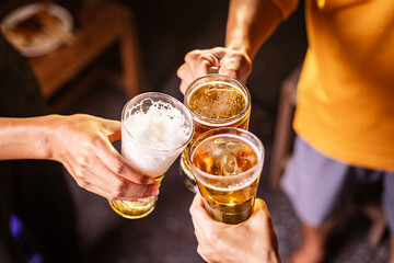 A man's hand and a woman's hand hold beer glasses in a cozy, lively evening party setting. Family and friends gather under the night sky, sharing joyful moments and laughter.