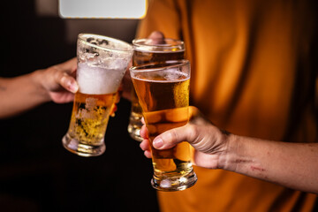 A man's hand and a woman's hand hold beer glasses in a cozy, lively evening party setting. Family and friends gather under the night sky, sharing joyful moments and laughter.