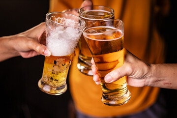 A man's hand and a woman's hand hold beer glasses in a cozy, lively evening party setting. Family and friends gather under the night sky, sharing joyful moments and laughter.