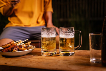 A man's hand and a woman's hand hold beer glasses in a cozy, lively evening party setting. Family and friends gather under the night sky, sharing joyful moments and laughter.