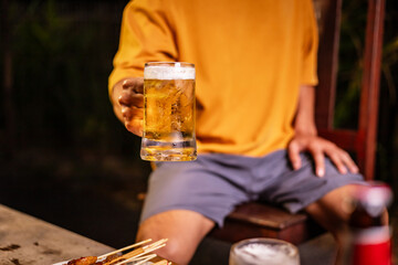 A man's hand and a woman's hand hold beer glasses in a cozy, lively evening party setting. Family and friends gather under the night sky, sharing joyful moments and laughter.