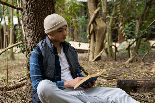 Young man reading book outdoors, relaxing against tree in peaceful forest