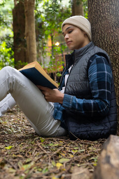 Young man reading book outdoors, relaxing against tree in peaceful forest