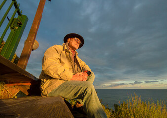 An old man in trench coat contemplating the sunset light at the seashore.