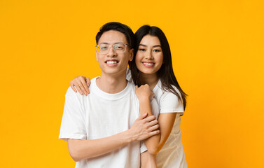 Naklejka premium Asian girl in love hugging her smiling boyfriend, yellow studio background