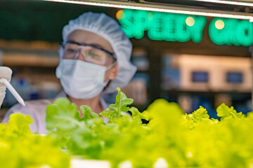 A scientist in a mask, goggles, and hairnet closely examines green plants in a lab setting. The...