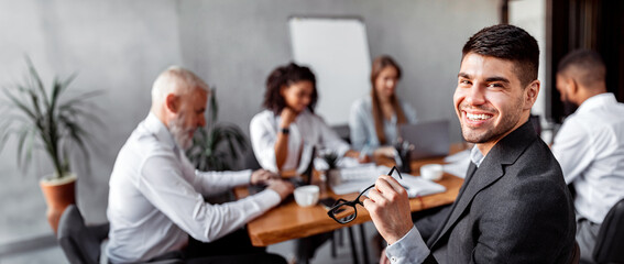 Young Businessman Smiling To Camera Sitting At Desk With Business Team During Corporate Meeting In Modern Office. Free Space
