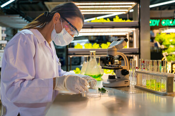 A scientist wearing a mask and gloves works in a lab, examining a green liquid in a beaker and using a microscope. The lab setup is well-lit, reflecting a focus on research, science, and experimentati