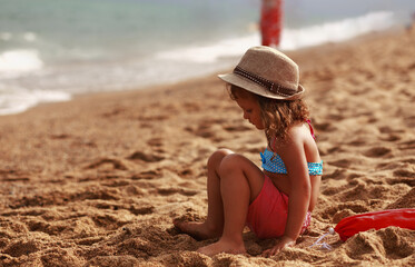 Beautiful fun happy kid girl thinking and sitting in summer clothing, straw hat on the yellow sand of the beach near blue sea and looking on water background. Happy