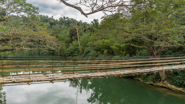 A rickety Bamboo Hanging Bridge spans over a tropical river. You can see the weaving of the footpath, the railing. Lush green vegetation on the shores. Reflection on calm water. Philippines.  Bohol 
