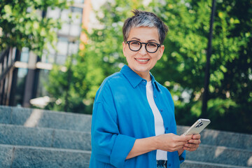 Portrait of pretty aged woman toothy smile hold smart phone wear blue shirt weekend walk city...