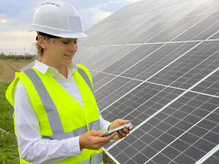 Young female engineer checks solar panels. A woman in a helmet and a vest with a phone in her hands...