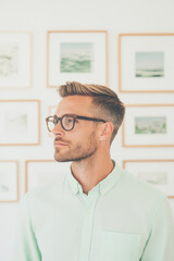 A stylish man with glasses stands thoughtfully against gallery wall adorned with framed photographs, exuding sense of calm and contemplation