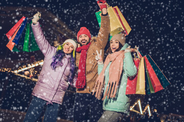 Photo of three excited overjoyed people hold shopping packages demonstrate v-sign enjoy tree garland lights outside