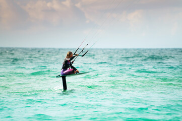 A young athlete in a colorful wetsuit sits on her hydrofoil and rides the waves with kitesurfing. Shot with a telephoto lens