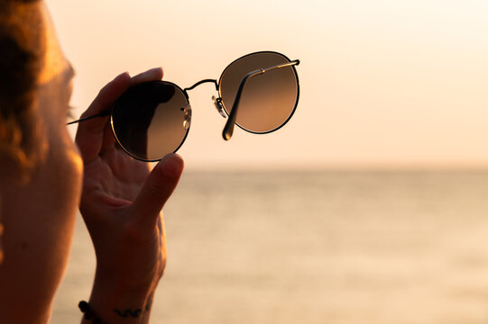 Close-up of a woman's hand holding sunglasses against the backdrop of the sea and sunset