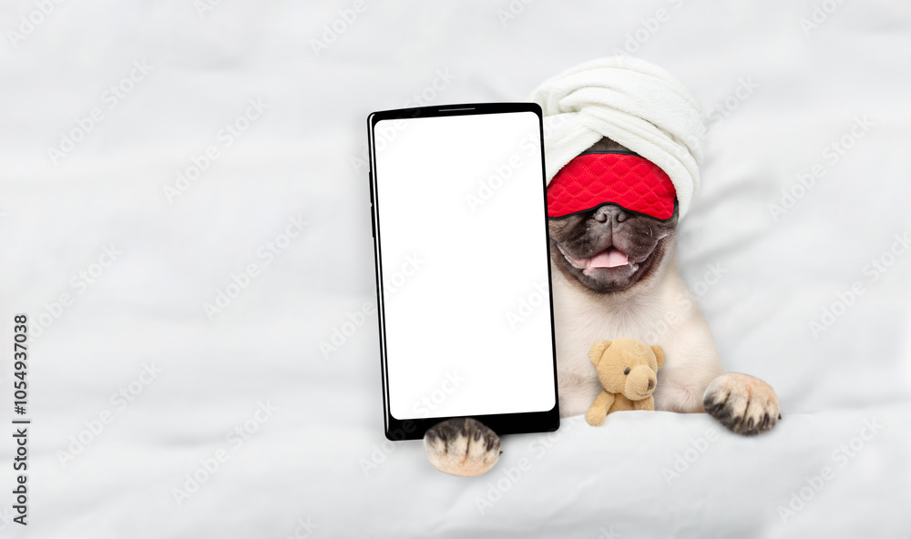 Poster Pug puppy wearing towel and sleeping mask lying with favorite toy bear on a bed at home and showing big smartphone with white blank screen in it paw. Top down view. Empty space for text