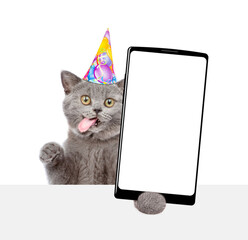 Happy kitten with tongue out wearing party cap shows big smartphone with blank screen above empty white banner. Empty space for text. isolated on white background
