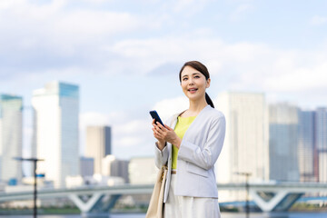 Fototapeta premium スマホ、スマートフォン、女性、ビジネスウーマン、ビジネス、キャリアウーマン、モバイル、5G、都市、通信、アプリ、屋外、青空、ブルー、社会、技術、テクノロジー、エレクトロニクス、携帯電話、ソーシャルメディア、ソーシャルネットワーク、SNS、ビル群、都会、笑顔、ユーザー、仕事、ビジネスマン、日本人、立つ、コピースペース、夕暮れ、夕焼け、タッチ、webサイト、インターネット、オンライン、メール、送信、人