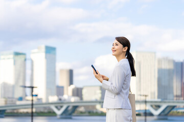 スマホ、スマートフォン、女性、ビジネスウーマン、ビジネス、キャリアウーマン、モバイル、5G、都市、通信、アプリ、屋外、青空、ブルー、社会、技術、テクノロジー、携帯電話、ソーシャルメディア、ソーシャルネットワーク、SNS、ビル群、都会、笑顔、ユーザー、仕事、ビジネスマン、日本人、立つ、コピースペース、webサイト、インターネット、オンライン、メール、人材、20代、30代、転職、就職、展望、将来、若い © metamorworks