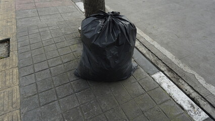 A black garbage bag that has been filled with garbage on the sidewalk during the day
