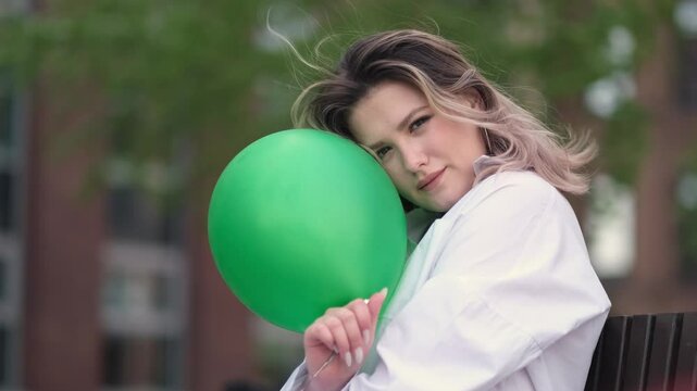 A young woman sits outdoors, a green balloon obscuring part of her face, masking her true expression. The playful setup contrasts with an underlying hint of sadness, reflecting themes of hidden