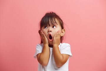 Amazed little asian girl in white t shirt shocked keeps hand on cheek, looks up, expresses...