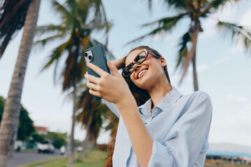 A woman in sunglasses takes a selfie with her phone, holding it in her palm, while standing in...