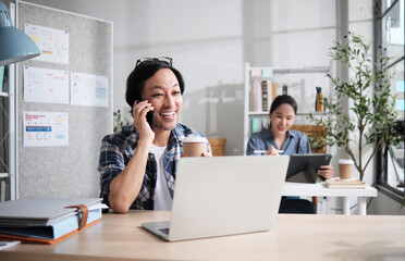 Asian startup partners working in a casual small business office, male worker talking on mobile phone with customers, online service jobs, female colleague behind, and E-commerce SME coworker team.