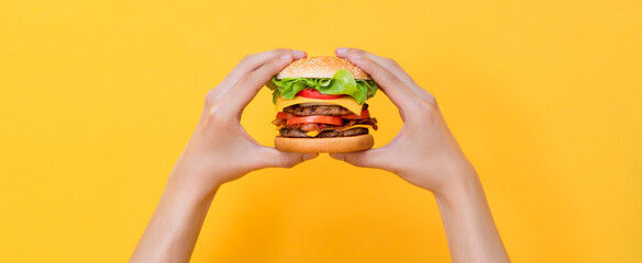 Hands holding fresh ready to eat hamburger, unhealthy and fast food concept, isolated on yellow color studio banner background