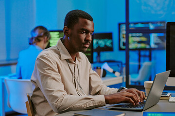 Medium shot of gen Z African American cybersecurity specialist typing on laptop at work in modern...
