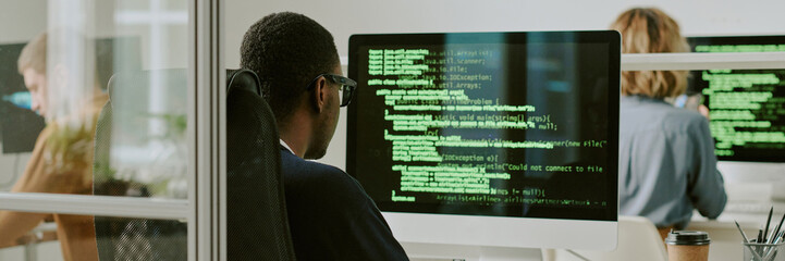Unrecognizable young Black man typing code on desktop computer at workplace in modern IT company office, website header