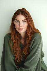 Fototapeta premium Portrait of a woman with long red hair wearing a green sweater against a neutral background, captured during daytime in a cozy indoor setting