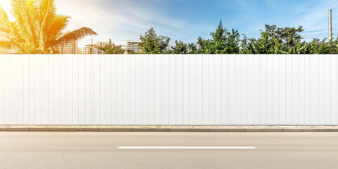 White fence separates lush tropical vegetation and city buildings from an asphalt road