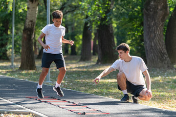 Obraz premium Boy exercises outdoors with a trainer using an agility ladder. The session promotes coordination, speed, and physical fitness in a fun, engaging environment under the guidance of a professional.