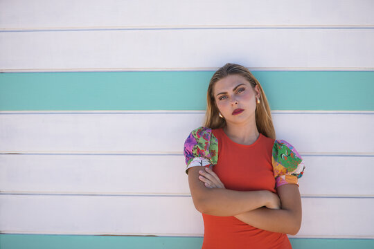 Young woman, beautiful, blue eyes, blonde, with orange colorful dress, arms crossed, empowered and independent leaning on a beach hut in the background. Concept beauty, fashion, trend, femininity. - Powered by Adobe