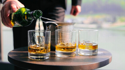 Bartender pouring whiskey into glasses with ice. A bartender's hand is seen pouring whiskey from a green bottle into glasses filled with ice cubes on a small round wooden table.