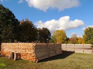 Stacked wooden planks lie on the grass against a backdrop of green and yellow trees and a blue sky...
