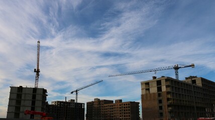 Three construction cranes sit behind unfinished multi-story buildings against a blue cloudy sky