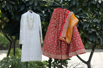 The image features a decorative display with traditional wedding attire. a richly embroidered lehenga and a cream-colored sherwani. placed against a vibrant red and gold grid backdrop.