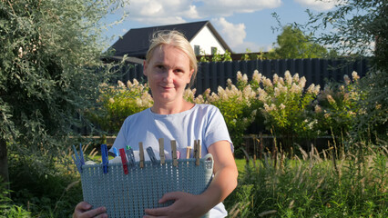 Middle-aged woman holding a basket filled with clothespins, standing in a lush garden on a sunny...