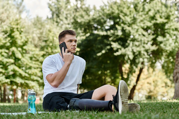 Fototapeta premium Tattooed young man with prosthetic leg sitting on fitness mat and having phone call, using smartphone in park