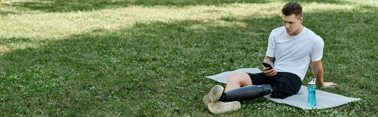 Tattooed young man with prosthetic leg sitting on fitness mat and using phone in green park