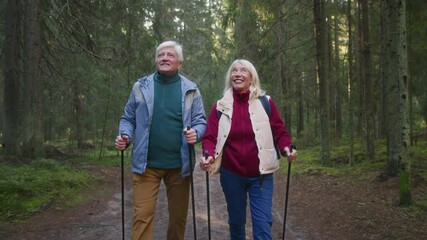 Senior couple enjoy Nordic walking with sticks on pathway in forest. Elderly positive man and woman stroll actively due to health caring. Energetic retirement marital pair smiling and admiring nature
