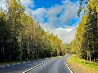 Peaceful scene of a winding road cutting through a lush forest in Karelia, with autumn leaves creating a warm, inviting atmosphere. Captured on a sunny day with a partly cloudy sky, this image