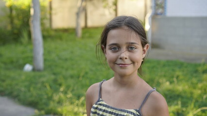 Close-up of a smiling young girl standing outside on a sunny day, with a green, blurred background suggesting a park or yard.